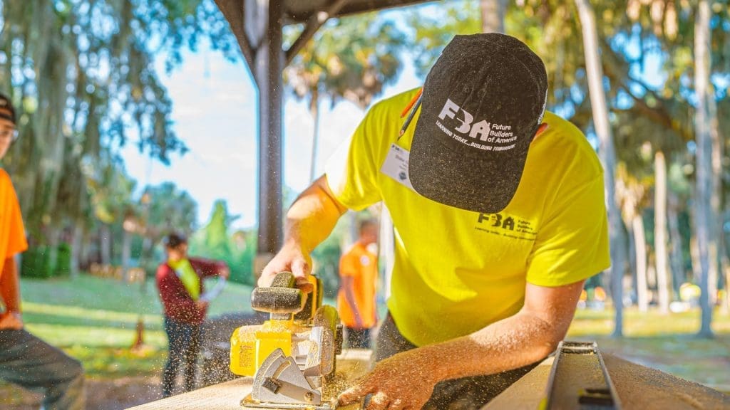 teenage man with navy bluehat with FBA logo on it sawing a piece of wood with woods in background.