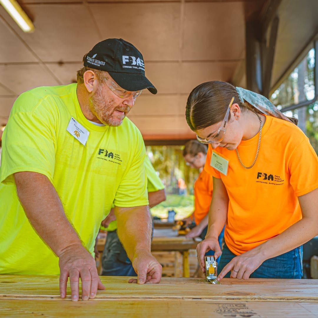 Instructor guiding a student during a hands-on woodworking activity at a Future Builders of America training program, both wearing safety glasses and FBA shirts.